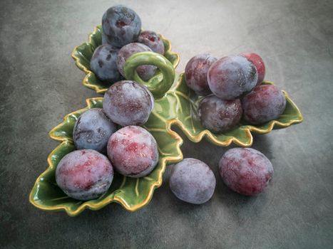 On the table in a ceramic dish are large ripe plums. Presented in close-up on a dark background.