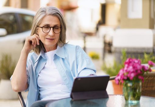 Charming european gray haired woman surf internet using digital tablet sitting at small street cafe. Mature silver hair woman working outdoors city cafe, wearing white tshirt and blue shirt.