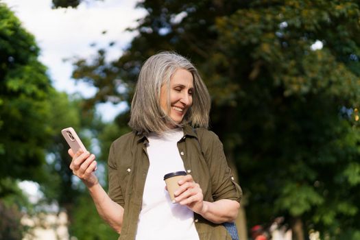 Mature european woman with gray hair happy enjoying free time after work or traveling holding phone while drinks coffee on the go using paper cup in city garden or park. Enjoying life mature woman.