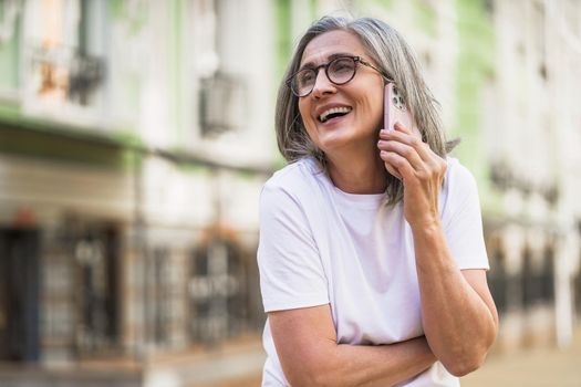 Charming mature business woman with grey hair talking on the phone holding smartphone standing outdoors of the streets of old urban city wearing white t-shirt. Silver hair woman outdoors.