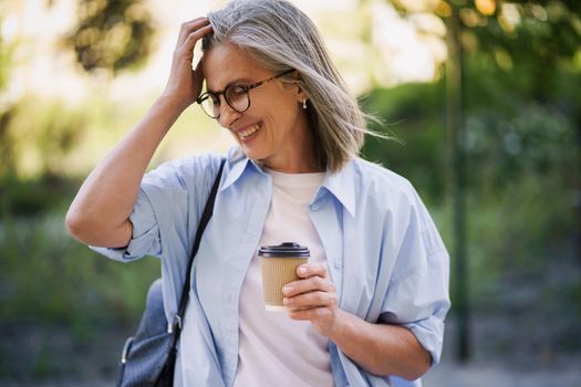 Charming mature woman drinks coffee on the go fixing her hair. Mature woman spend free time traveling in european old town enjoying cup of tea, coffee. Grey haired woman in casual with backpack.