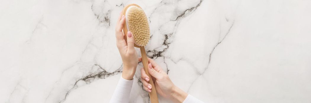 Brush for dry massage in the hands of a girl on a marble background. Brush for scrubbing the body, bringing it in order, against cellulite. Web banner.