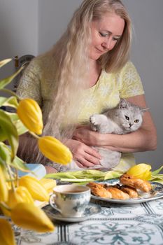 blonde middle-aged hugging a kitten and drinking tea, still life with yellow tulips and cakes a stack of books on the table. High quality photo