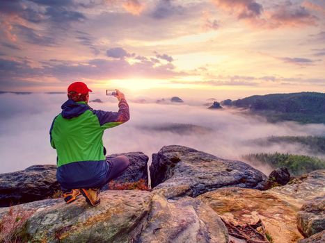 Hiker takes selfie photo, fall nature adventure. Man sit on stone on mountain sumit  Daybreak horizon above milky fog.