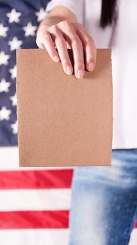 Young woman holds empty cardboard with Space for Text sign against American flag on background. Girl protesting anti-abortion laws. Feminist power. Womens rights freedom