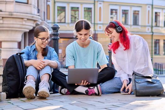 Teenage friends in headphones looking at laptop. Group of young people having fun laughing looking at laptop screen, sitting on sidewalk, urban style background. Youth, fun, lifestyle, leisure concept