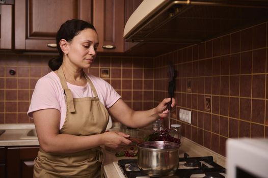 Multiethnic middle-aged woman confectioner, a housewife wearing a chef apron, standing by stove and cooking cherry jam in the pot at home wooden kitchen