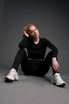 Full body of positive young athletic woman with long ginger hair in stylish sportswear and sneakers sitting on floor and looking at camera in gray studio