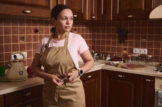Charming middle-aged multiethnic woman, housewife, chef confectioner putting on a chef apron in a cozy rustic home wooden kitchen interior.