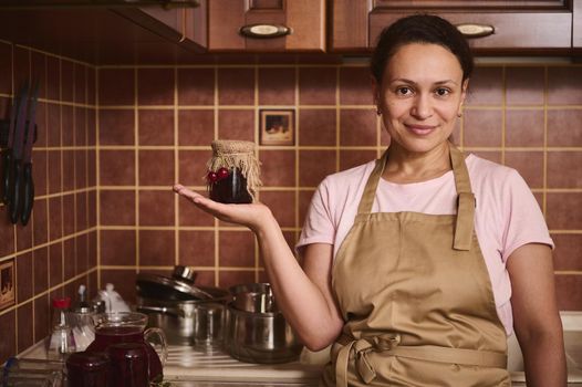 Pleasant charming Middle-Eastern woman confectioner, housewife in chef apron holds a jar of homemade confiture on her hand palm up and smiles, looking at camera, standing in a cozy rustic home kitchen