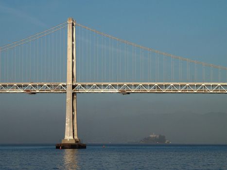 Sun shines on the Bay Bridge with Alcatraz island in the distance with a lite layer of fog.