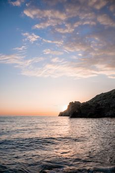 A red burning sunset with the silhouette of a cliff over the sea. Abstract nature summer or spring ocean sea background. Small waves on golden warm water surface with bokeh lights from sun.