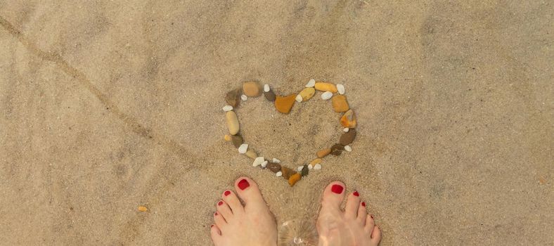 banner with women's feet on the beach and a heart is laid out of pebbles. relaxing on the beach, summer by the sea