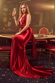 Young blond girl in a long red satin dress, with a glass of champagne in her hand is posing sideways sitting on a poker table in luxury casino. Passion, cards, chips, alcohol, win, gambling - it is a female entertainment. Smoke background.