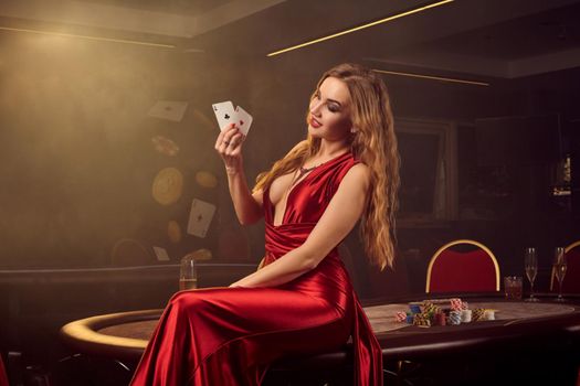 Showy blond maiden in a long red satin dress, with two aces in her hand is posing sitting sideways on a poker table in luxury casino. Passion, cards, chips, alcohol, win, gambling - it is a female entertainment. Smoke background.