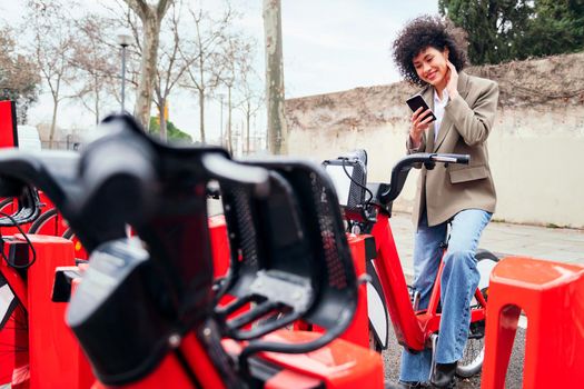 smiling young woman checking her phone while sitting on a bike at the rental station, technology and sustainable mobility concept