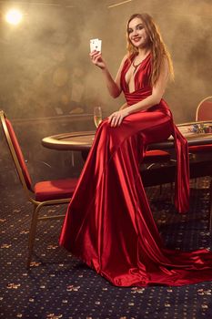 Young blond maiden in a long red satin dress, with two aces in her hand is smiling widely and posing sitting on a poker table in luxury casino. Passion, cards, chips, alcohol, win, gambling - it is a female entertainment. Smoke background.