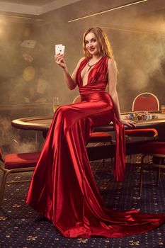 Young blond lady in a long red satin dress, with two aces in her hand is smiling and posing sitting on a poker table in luxury casino. Passion, cards, chips, alcohol, win, gambling - it is a female entertainment. Smoke background.