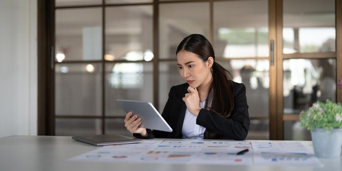 Portrait seriously business asian woman using digital tablet for do math analysis finance on wooden desk in office, tax, accounting, financial concept.