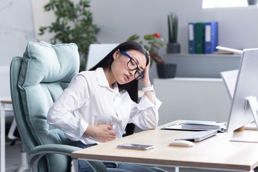 Abdominal pain at the workplace. A young beautiful Asian woman in glasses works in the office at the computer, feels pain in her stomach, holds her hand to her stomach, grimaced in pain.