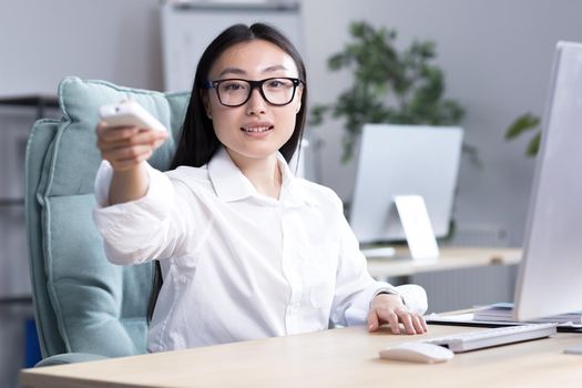 Heat in the workplace. A young Asian business woman in glasses, a white shirt sits in the office at the desk, turns on the air conditioner with the remote control, needs fresh air.