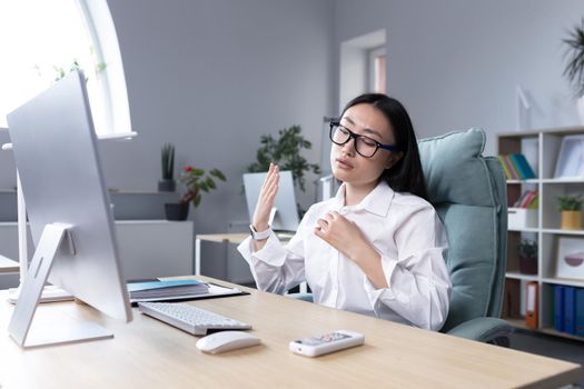 Heat in the workplace. Young Asian woman sitting in the office at the desk waving her hand, it's very hot, she feels bad, she needs fresh air, take a break.