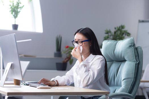 Feeling unwell at the workplace.A young Asian business woman, freelancer, manager, employee fell ill. He is sitting at the desk wiping his nose with a napkin, has a runny nose, and feels bad.