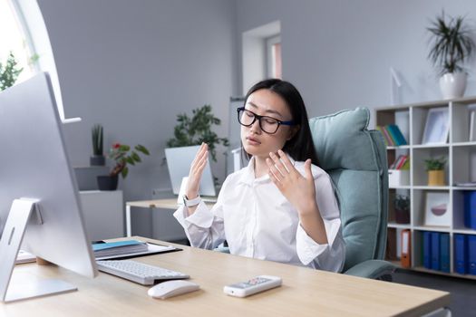 Heat in the workplace. A young Asian woman sits in the office at the desk, waves both hands at her face, it is very hot, she feels bad, she needs fresh air, take a break.