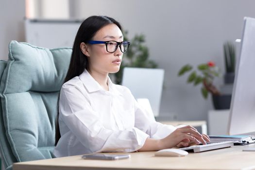 Young Asian girl student in glasses sits and studies at the computer online remotely at home at the table, typing on the keyboard, passing the exam, serious.