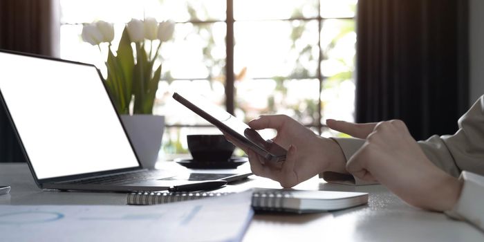 Cropped shot of female blogger searches information on internet websites, uses portable laptop with blank copy screen for your advertisment. Modern cell phone in woman`s hands, selective focus.