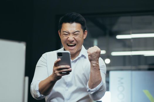 Close-up photo portrait of successful Asian businessman triumphantly reading news from phone, celebrating triumph success and victory, holding hand up