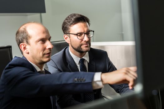 Image of two thoughtful businessmen looking at data on multiple computer screens, solving business issue at business meeting in modern corporate office. Business success concept.