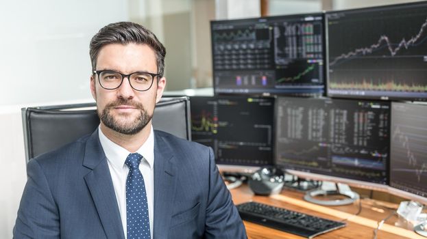 Business portrait of confident stocks broker holding a business newspaper in traiding office with multiple computer screens full of index charts and data analyses.