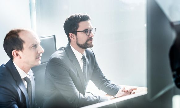 Image of two thoughtful businessmen looking at data on multiple computer screens, solving business issue at business meeting in modern corporate office. Business success concept.