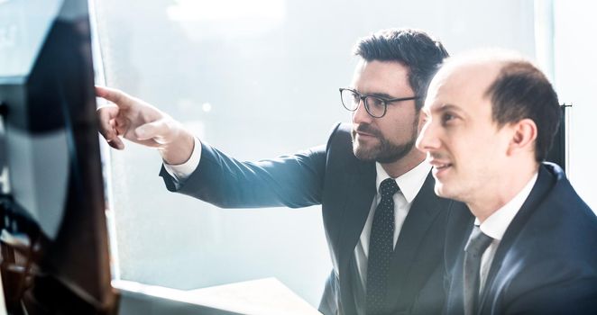 Image of two thoughtful businessmen looking at data on multiple computer screens, solving business issue at business meeting in modern corporate office. Business success concept.