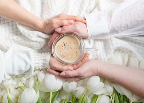 four hands hold one cup of heart-shaped cappuccino, floral background with white tulips, drink coffee in the company, female friendship, lgbt. High quality photo