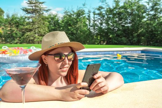 young red-haired woman relaxing by the pool, drinking a cocktail and looking at her smartphone. girl on holiday sharing the moments on her social networks. summer concept and free time. outdoor garden, natural sunlight.