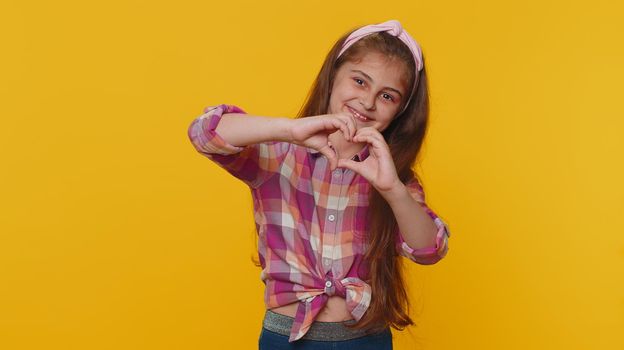 I love you. Smiling young preteen child girl kid makes heart gesture demonstrates love sign expresses good feelings and sympathy. Little toddler children isolated alone on studio yellow background