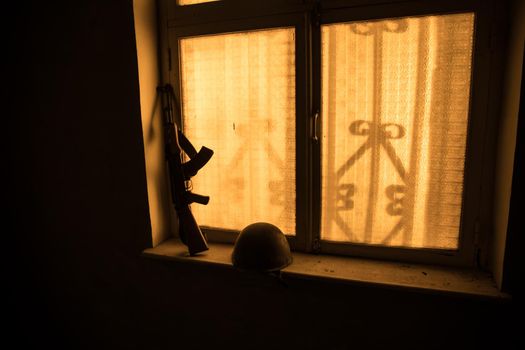 Conceptual photo of war between Russia and Ukraine. Russian weapon and helmet on windowsill at night. Old creepy room with window. Explosion outside.