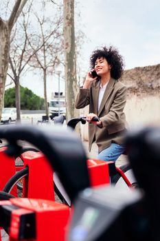 smiling young latin woman talking by phone while sitting on a bike at the rental station, technology and sustainable mobility concept