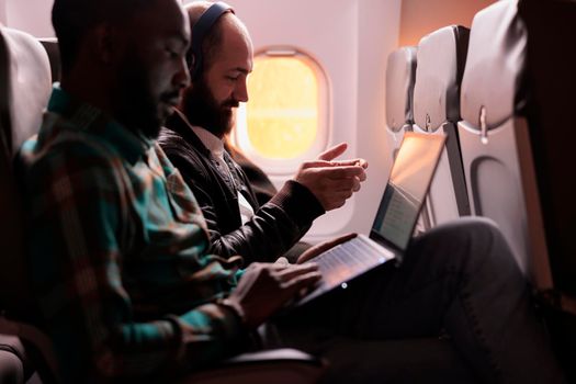 Diverse group of travellers flying abroad in economy class, using laptop, smartphone and headphones during sunset flight. Passengers travelling with international commercial airline.