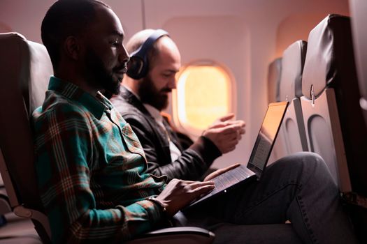 Young freelancer travelling on work trip by airplane in economy class, working on laptop during sunset flight. Male employee flying with commercial flight with international airline.