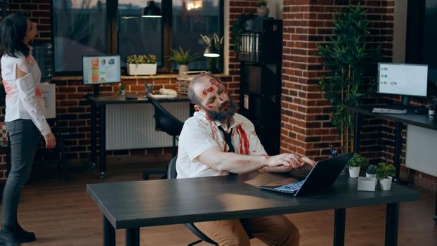 Scary zombie sitting in office while working on modern laptop. Mindless messy brain dead monster with deep and bloody wounds sitting at table in workspace while trying to use portable computer