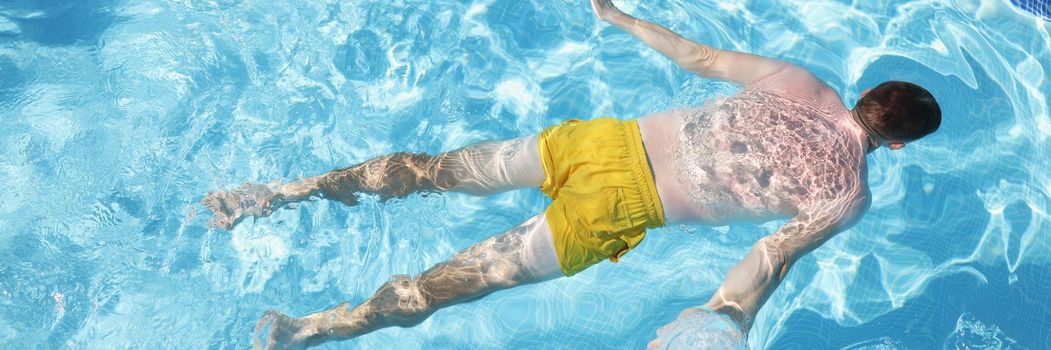 Top view of male person in swimming pants floating on water on belly, clear water in pool. Man relax and sunbathe under summer sun. Holiday, resort concept