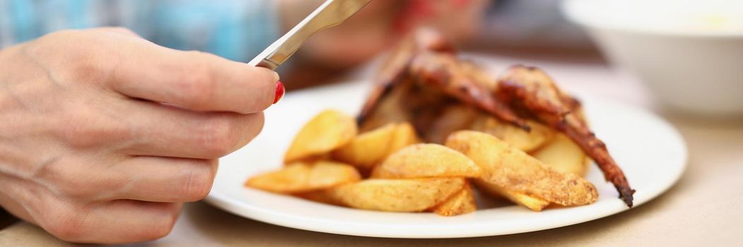 Close-up of person having breakfast or lunch in cafe or restaurant, hold knife and fork. Chicken wings and fried potatoes on plate. Food, hunger concept