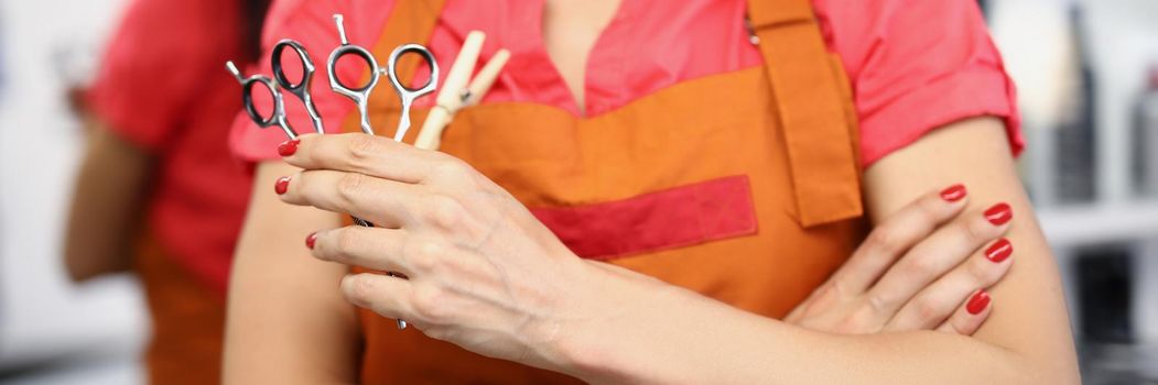 Close-up of young professional master holding equipment for work and posing in uniform. Qualified hairdresser hold pair of scissors. Beauty studio concept