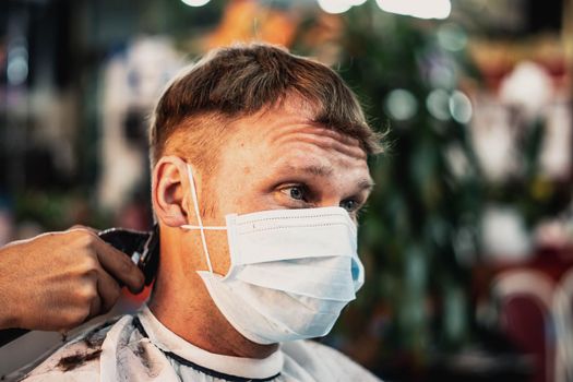 Lifestyle photo barbershop. Young man customer wears face mask, surprised dissatisfied look, wrinkled forehead. Hairdresser male hand work comb tool electric clipper. Short hair locks cut.