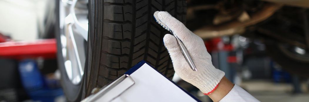 Close-up of mechanic worker point at tyre and write down in document notes. Examine damaged car, add adjustments. Car service, handyman, pit stop concept