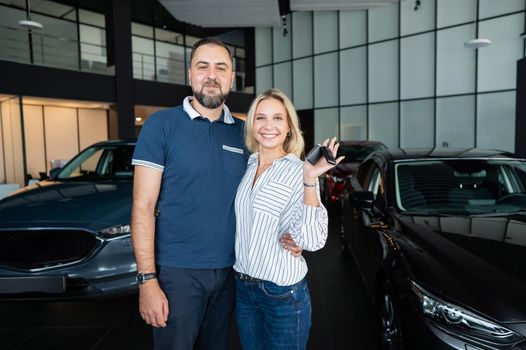 Happy caucasian couple hugging and woman holding new car keys in car showroom