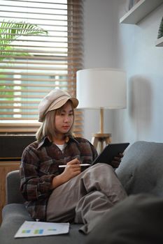 Young asian woman siting on sofa and analyzing stock market trading on digital tablet.